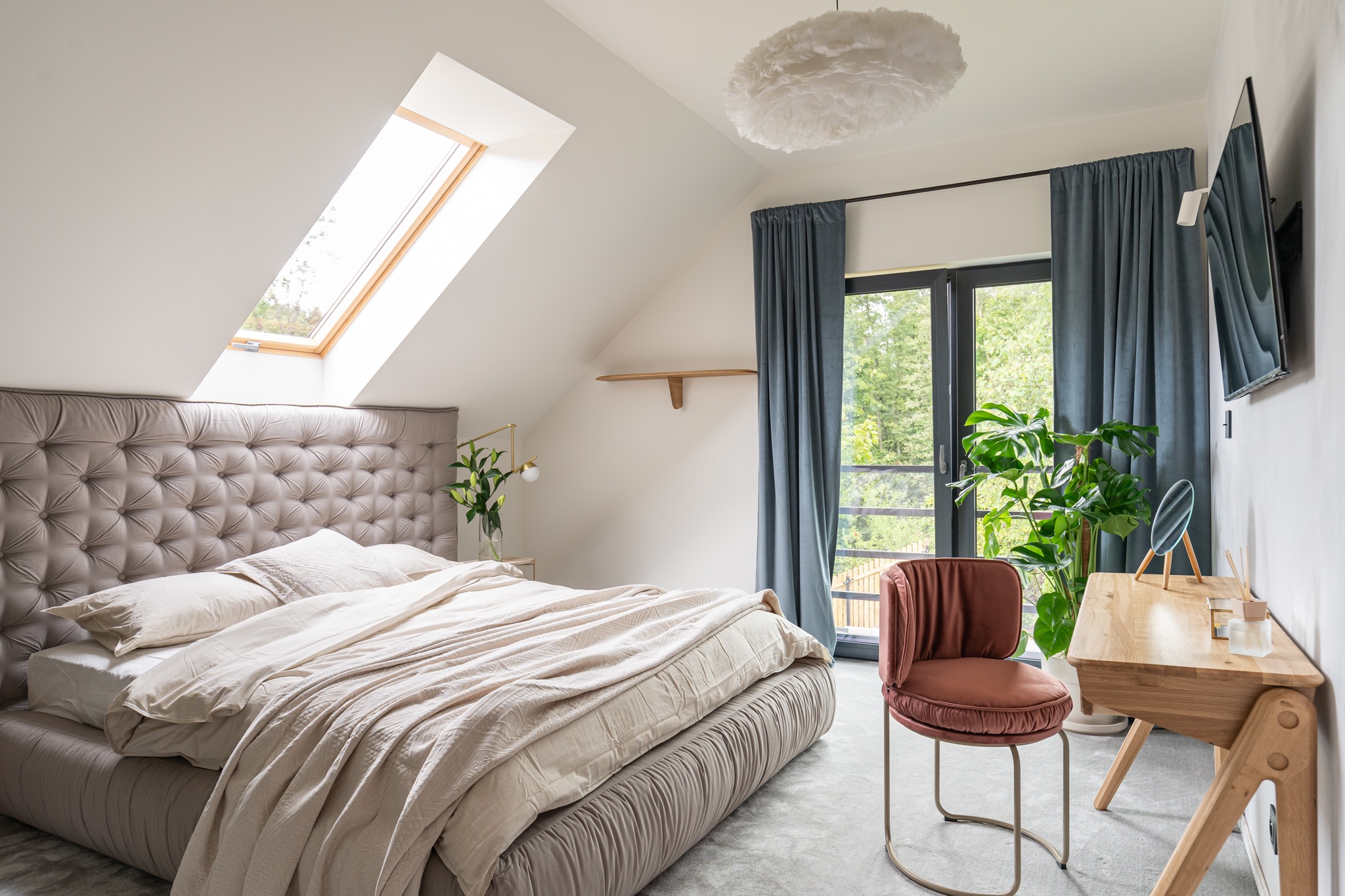 Jasna sypialnia na poddaszu z łóżkiem z pikowanym zagłówkiem, oknem dachowym, biurkiem z lustrem i welurowym krzesłem w kolorze terakoty na tle okna balkonowego z zasłonami.