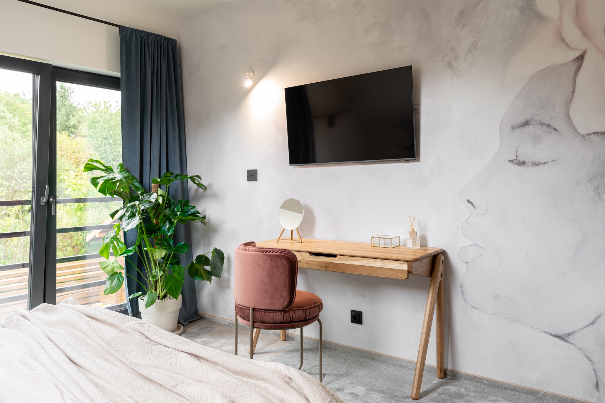 Nowoczesna sypialnia z biurkiem z jasnego drewna, różowym krzesłem, telewizorem na ścianie z artystycznym malunkiem, dużą rośliną doniczkową przy oknie i beżową narzutą na łóżku.