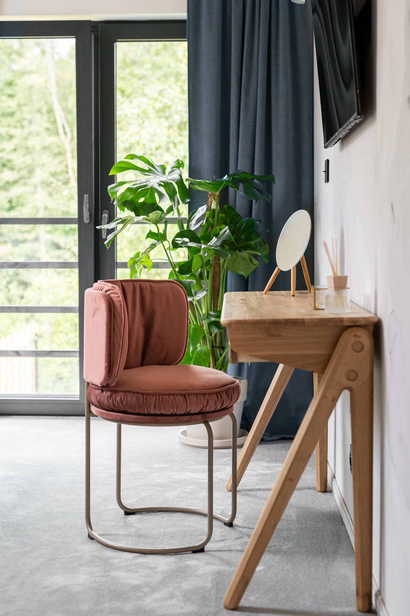 Nowoczesne wnętrze z pudrowo-różowym krzesłem welurowym, drewnianym biurkiem, lustrem na nóżkach i rośliną Monstera w tle, oświetlone przez okno z ciemną zasłoną.