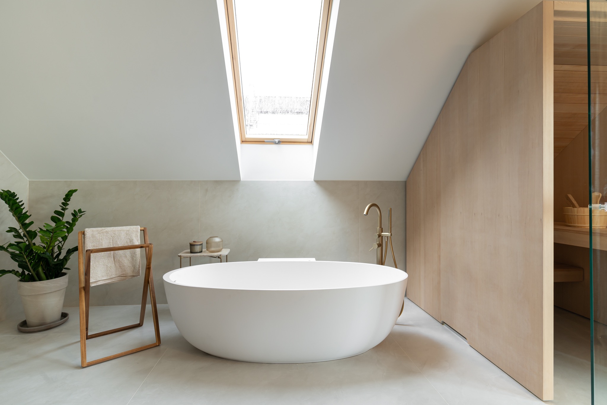 Nowoczesna łazienka na poddaszu z wolnostojącą białą wanną, złotą armaturą, drewnianą ścianą i sauną, oświetlona przez okno dachowe, z rośliną doniczkową i wieszakiem na ręczniki.