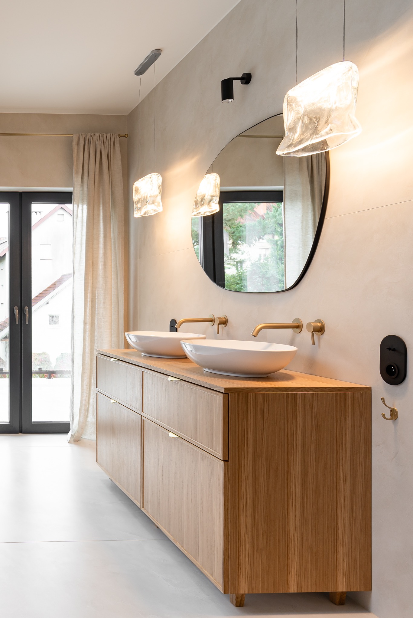 Nowoczesna łazienka z dwiema owalnymi umywalkami nablatowymi, drewniana szafka z subtelnymi uchwytami, okrągłe lustro z czarną ramą i designerskie lampy wiszące o nieregularnym kształcie, widok...