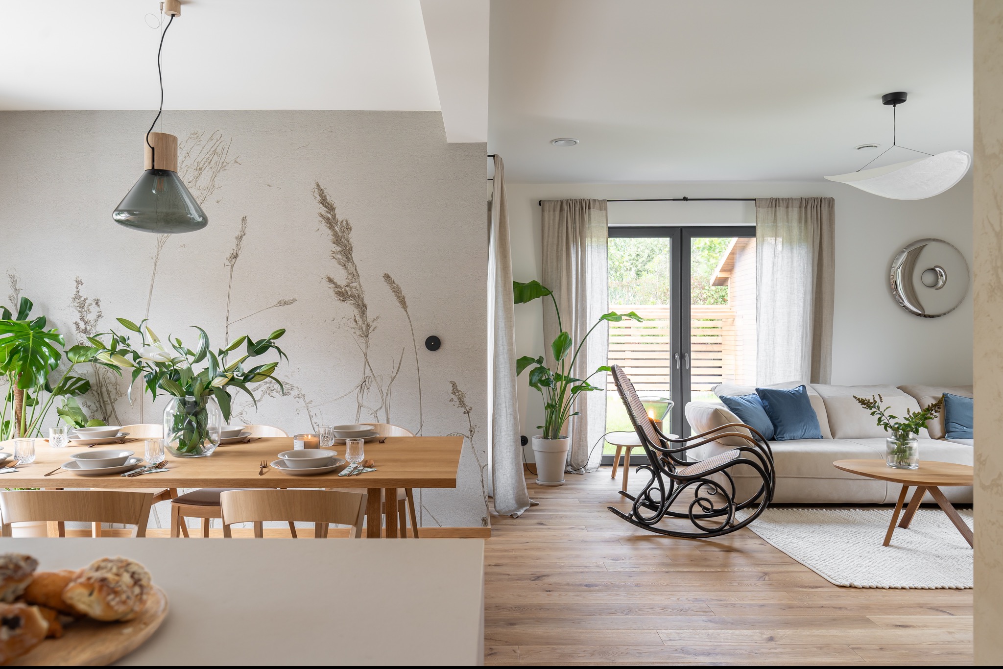 Jasne, przestronne wnętrze z drewnianym stołem jadalnym nakrytym zastawą, kwiatami w wazonie i modernistyczną lampą, połączone z salonem z wiklinowym fotelem bujanym, kremową sofą z niebieskimi...