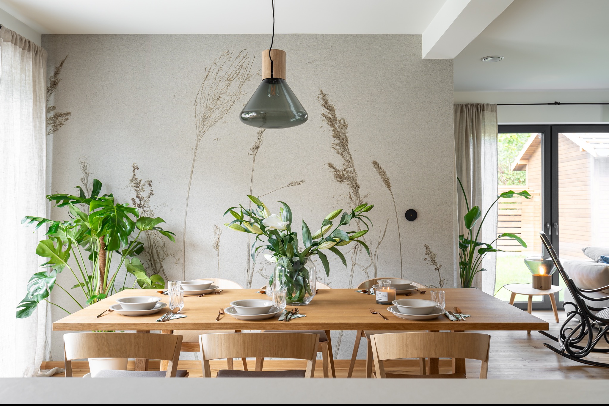 Jadalnia w neutralnych barwach z drewnianym stołem, nakrytym do posiłku, z dekoracyjnym wazonem z liliami i tapetą w motywy traw. W tle widoczny salon z bujanym fotelem.