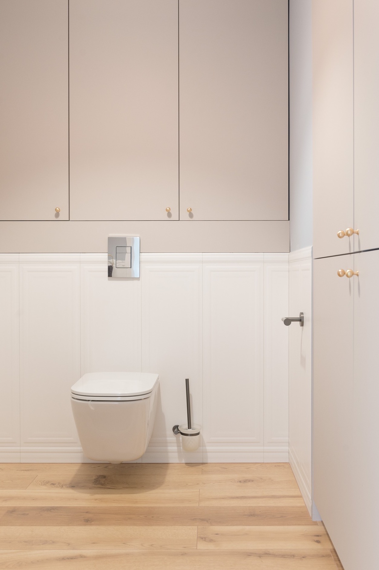 Nowoczesna łazienka z podwieszaną toaletą, białą boazerią ścienną, beżowymi szafkami z okrągłymi złotymi uchwytami i drewnianą podłogą.