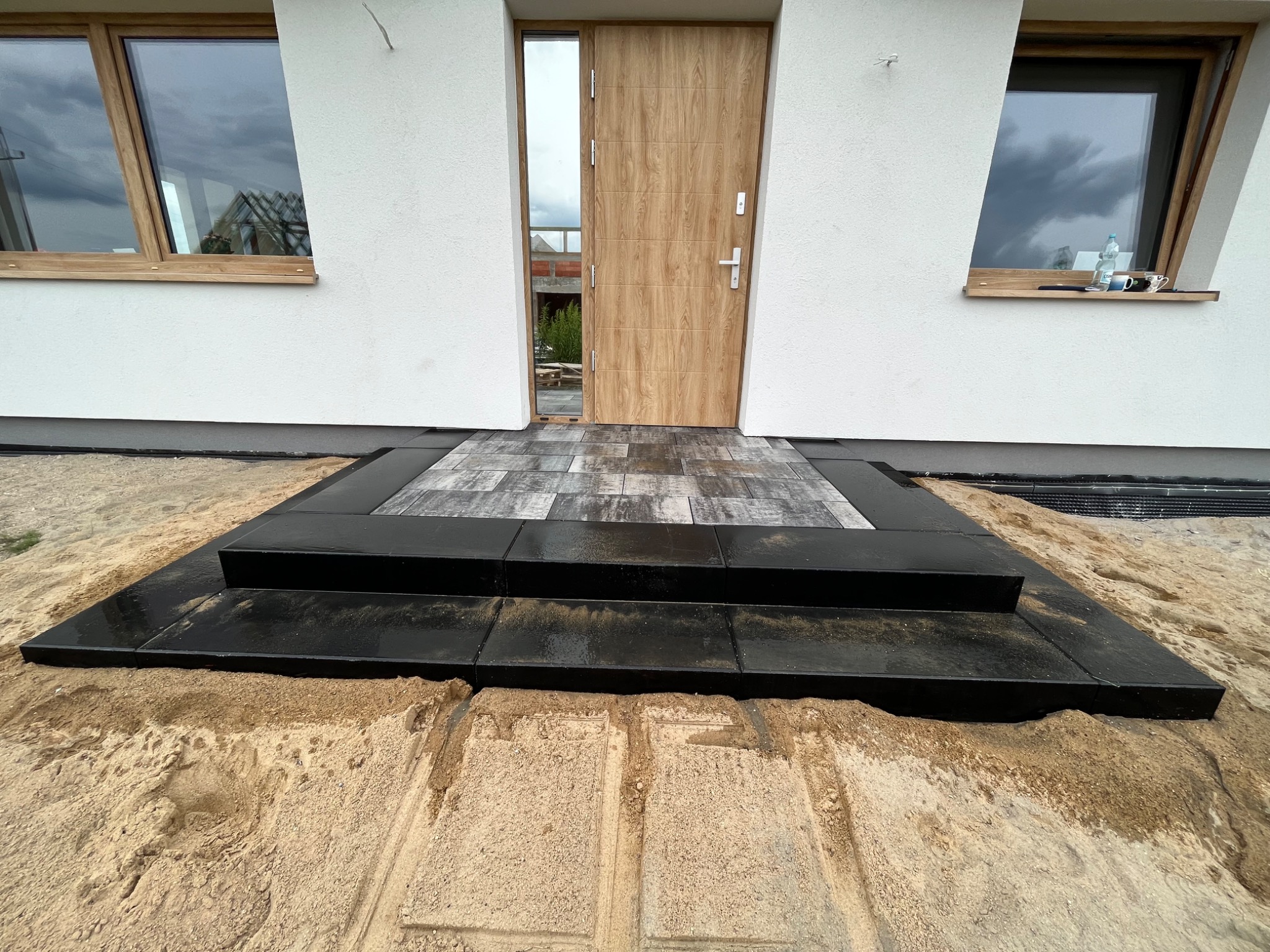 Schody wejściowe do domu, wykończone czarną kostką brukową i szarą płytą na górnym stopniu, otoczone piaskiem, z widokiem na białą elewację budynku i drewniane okna.