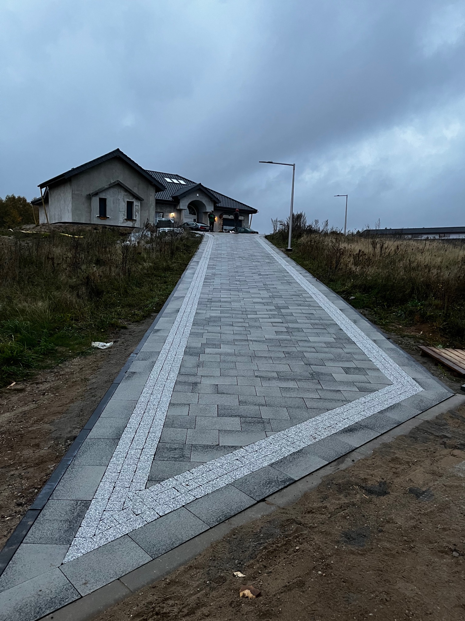 Nowo ułożona, długa, szara aleja z kostki brukowej prowadząca do domu w stanie surowym zamkniętym, z dekoracyjnym, jasnym wzorem w kształcie litery V. Brzegi wykończone ciemnoszarą kostką.