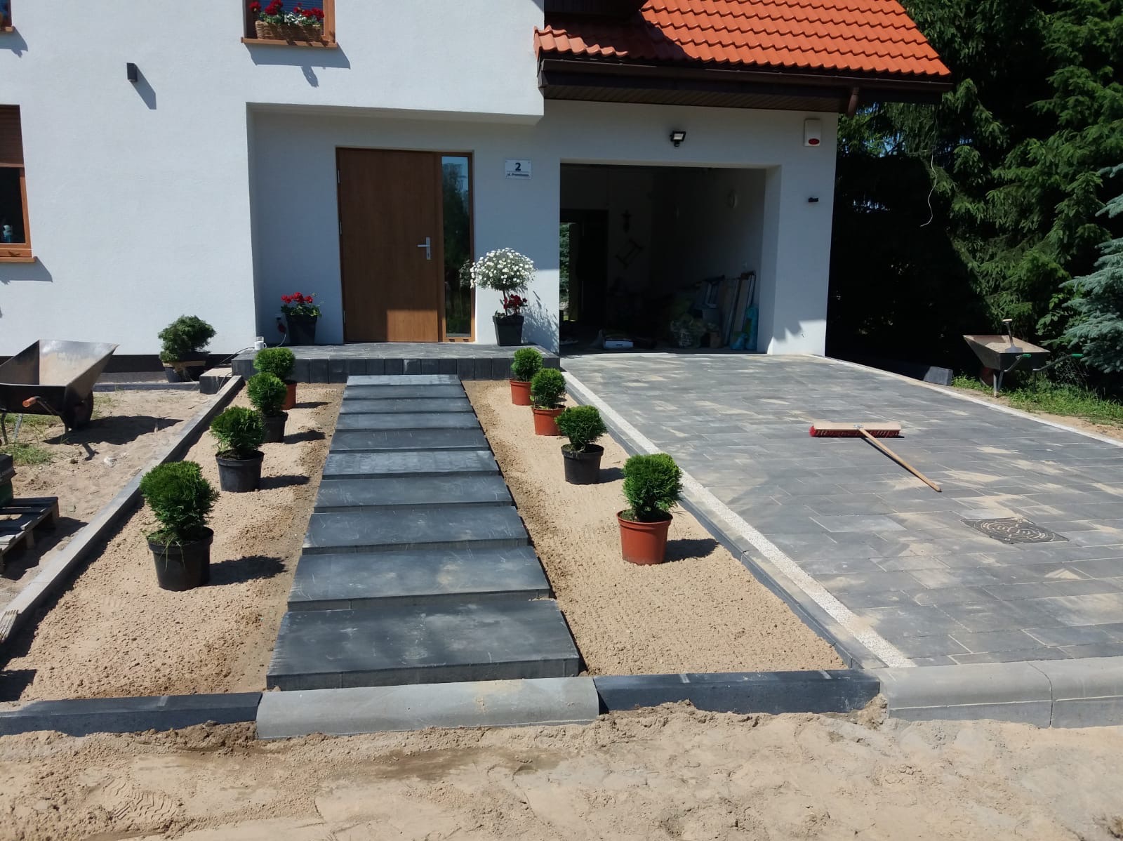 Nowoczesne podejście do domu z ciemnoszarych płyt chodnikowych, otoczone piaskiem i niskimi krzewami w donicach, prowadzące do drzwi wejściowych i garażu z podjazdem z kostki brukowej. Równiarka...