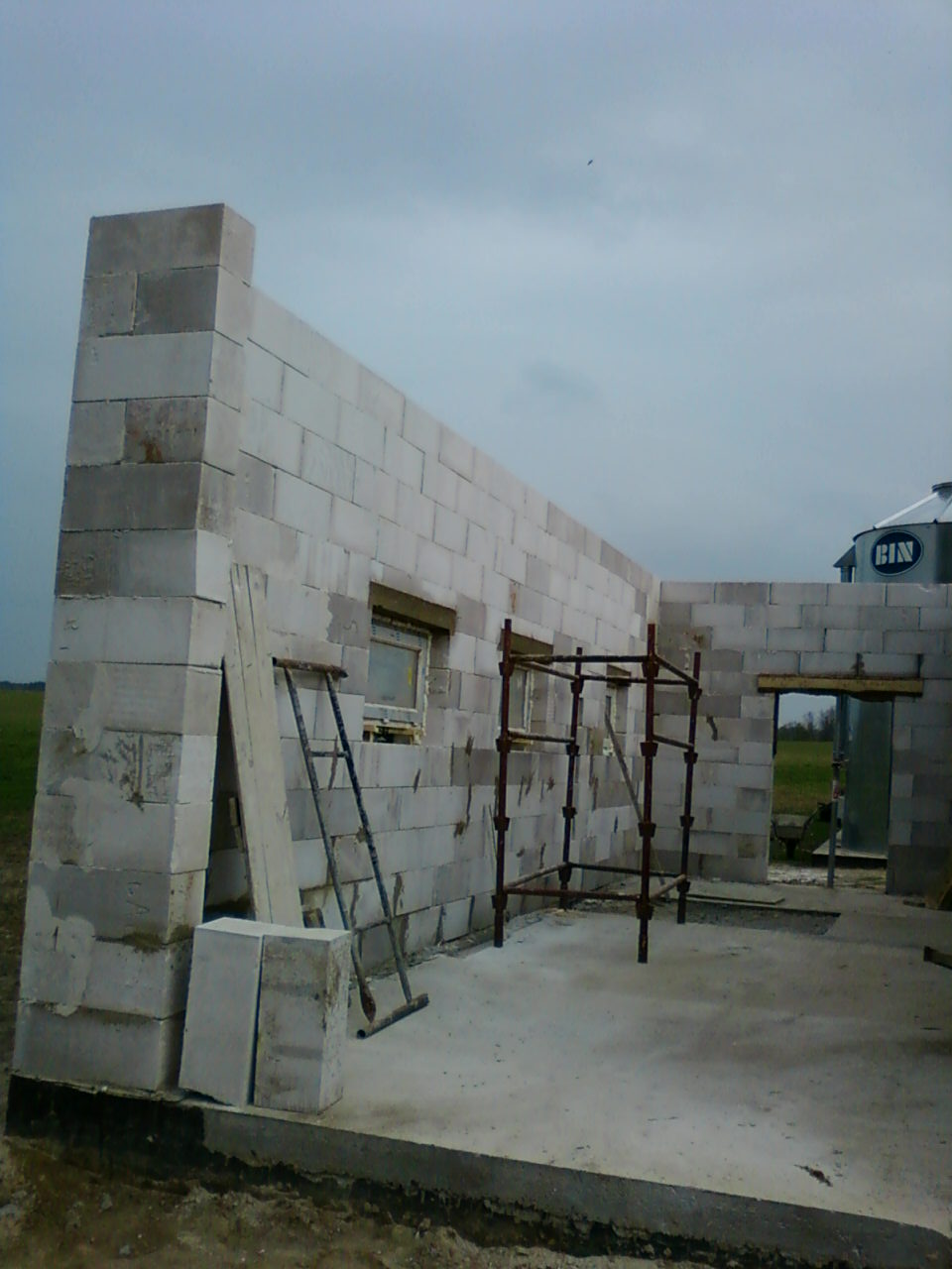 Wznoszenie ścian budynku z betonu komórkowego, widoczne rusztowanie i podłoże betonowe, w tle silos zbożowy.