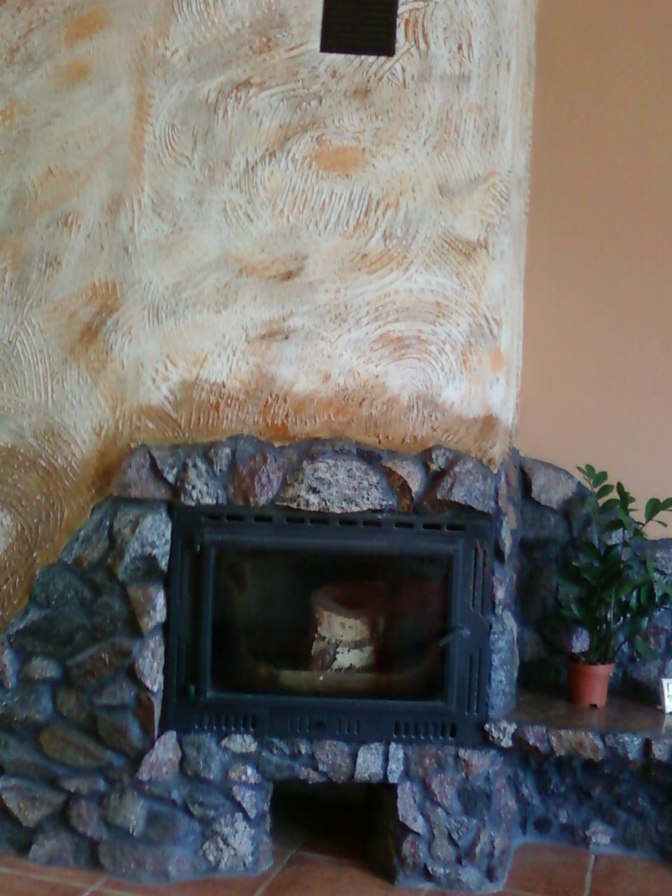 Ozdobny kominek z kamienną obudową i żeliwnym wkładem, ściana z dekoracyjnym tynkiem, obok doniczka z rośliną.