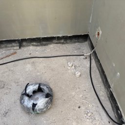 Systemele - Wnętrze w trakcie prac elektrycznych: kabel w peszlu prowadzony wzdłuż ściany, puszki elektryczne z przewodami, zwój kabla w folii na podłodze. Surowy stan wykończenia.