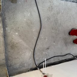 Systemele - Czarny kabel elektryczny wychodzący z otworu w podłodze, białe przewody obok. Fragment podłogi z wylewką i poziomica w tle. Prace instalacyjne w trakcie.