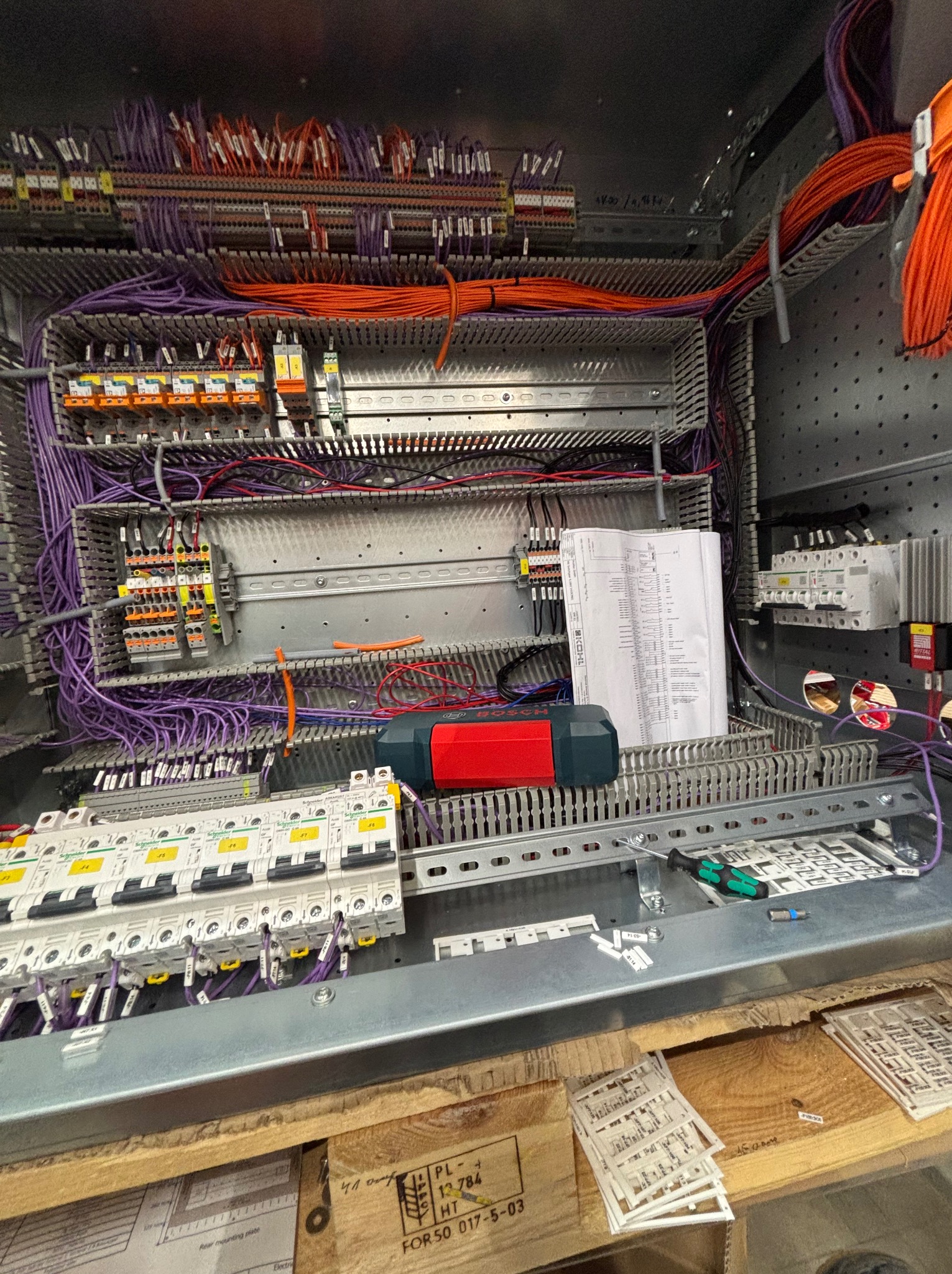Rozdzielnia elektryczna w trakcie montażu: liczne przewody (pomarańczowe, fioletowe), bezpieczniki, listwy zaciskowe, narzędzia i dokumentacja techniczna wewnątrz metalowej obudowy.
