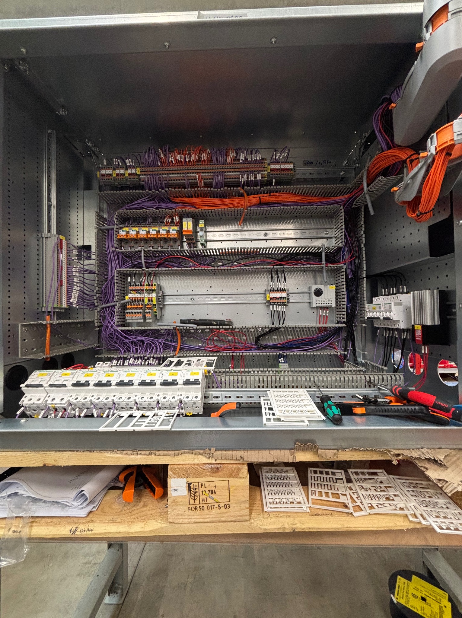 Rozbudowana szafa sterownicza w trakcie montażu. Widoczne liczne przewody, bezpieczniki, listwy zaciskowe i narzędzia na drewnianej podstawie. Skrzynka drewniana z oznaczeniami.