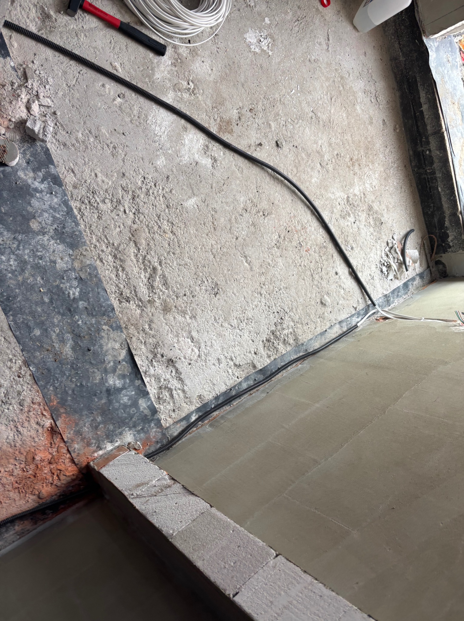 Wnętrze w trakcie prac: kable elektryczne w peszlu ułożone wzdłuż ściany, częściowo zalane wylewką. Widoczne narzędzia i przewody. Ujęcie z góry, ukośne.