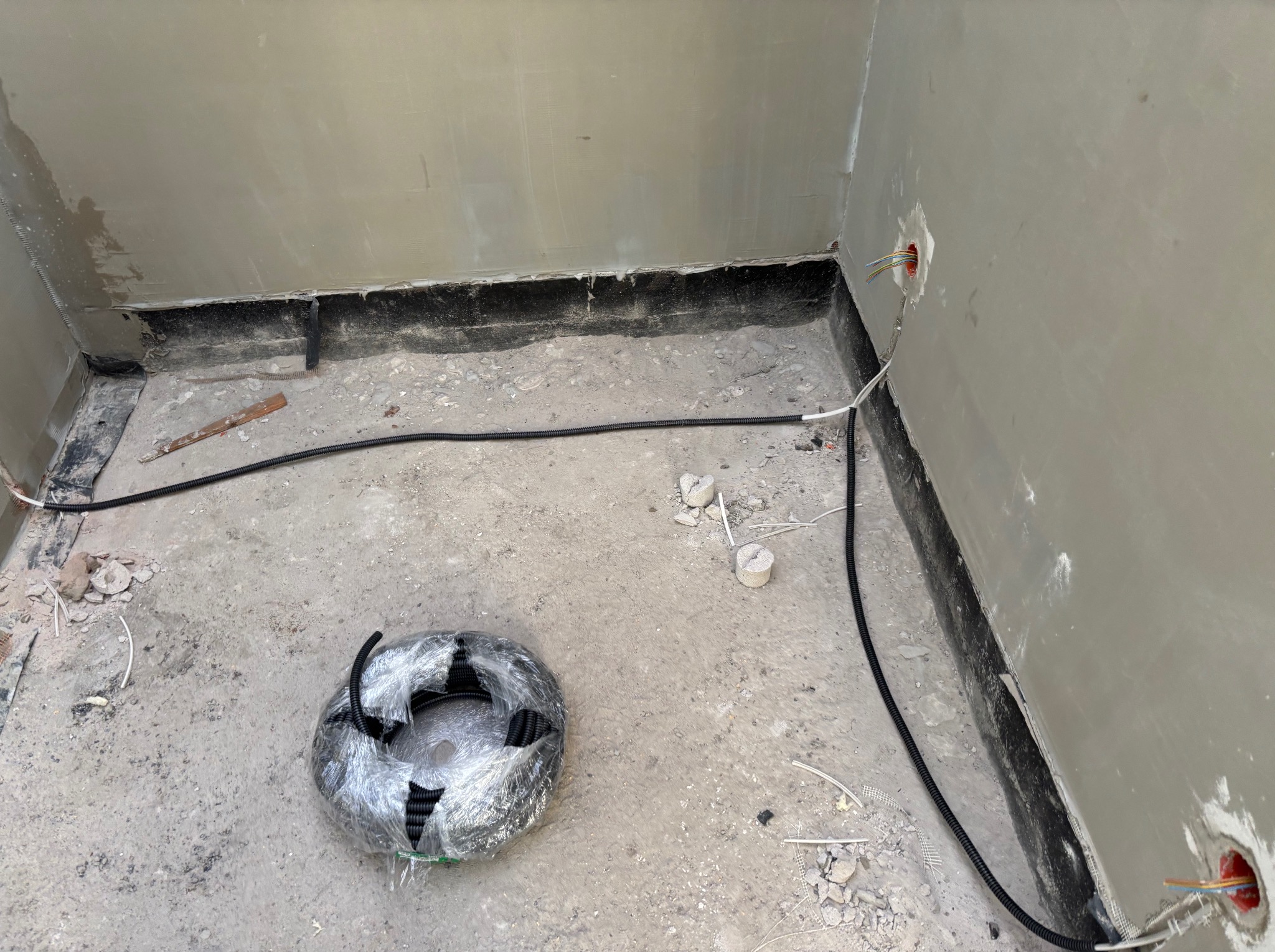 Wnętrze w trakcie prac elektrycznych: kabel w peszlu prowadzony wzdłuż ściany, puszki elektryczne z przewodami, zwój kabla w folii na podłodze. Surowy stan wykończenia.