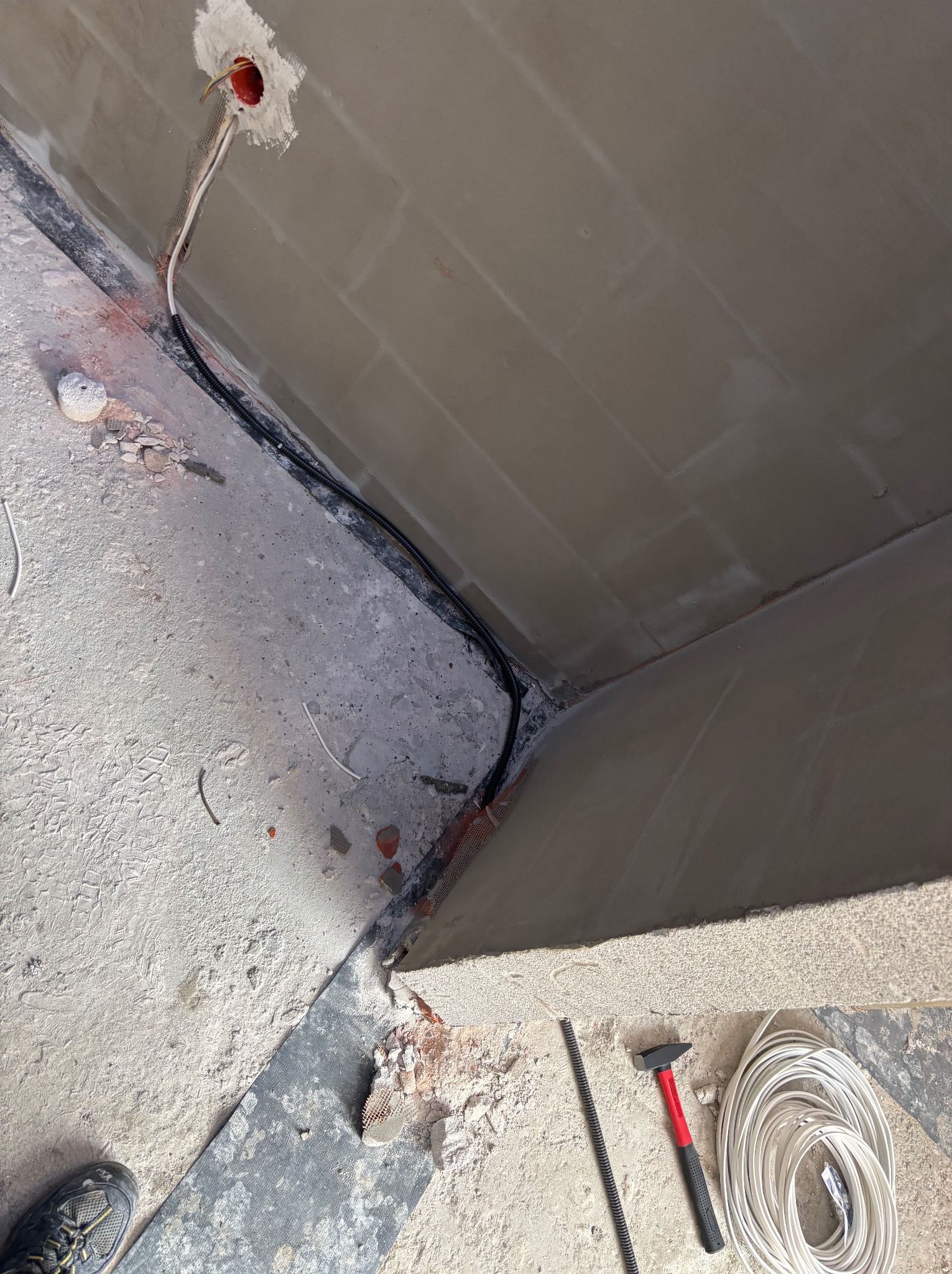 Fragment wnętrza w trakcie prac elektrycznych: przewody w ścianie, puszka instalacyjna, narzędzia (młotek, zwój kabla). Widoczne ślady prac budowlanych.