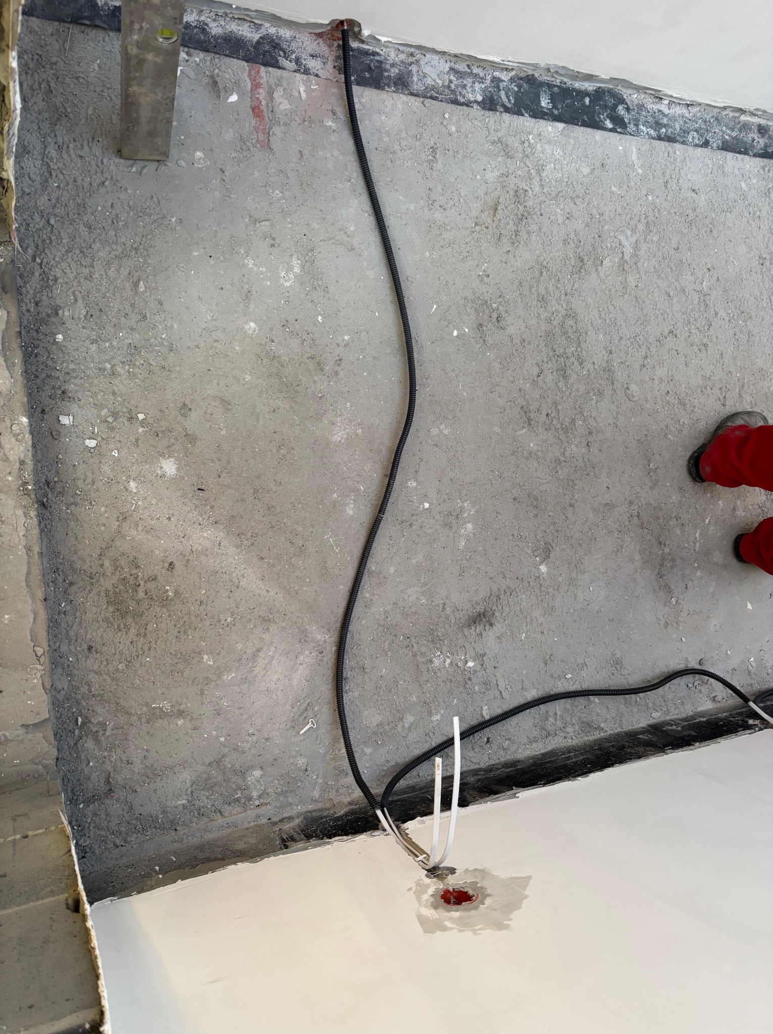 Czarny kabel elektryczny wychodzący z otworu w podłodze, białe przewody obok. Fragment podłogi z wylewką i poziomica w tle. Prace instalacyjne w trakcie.