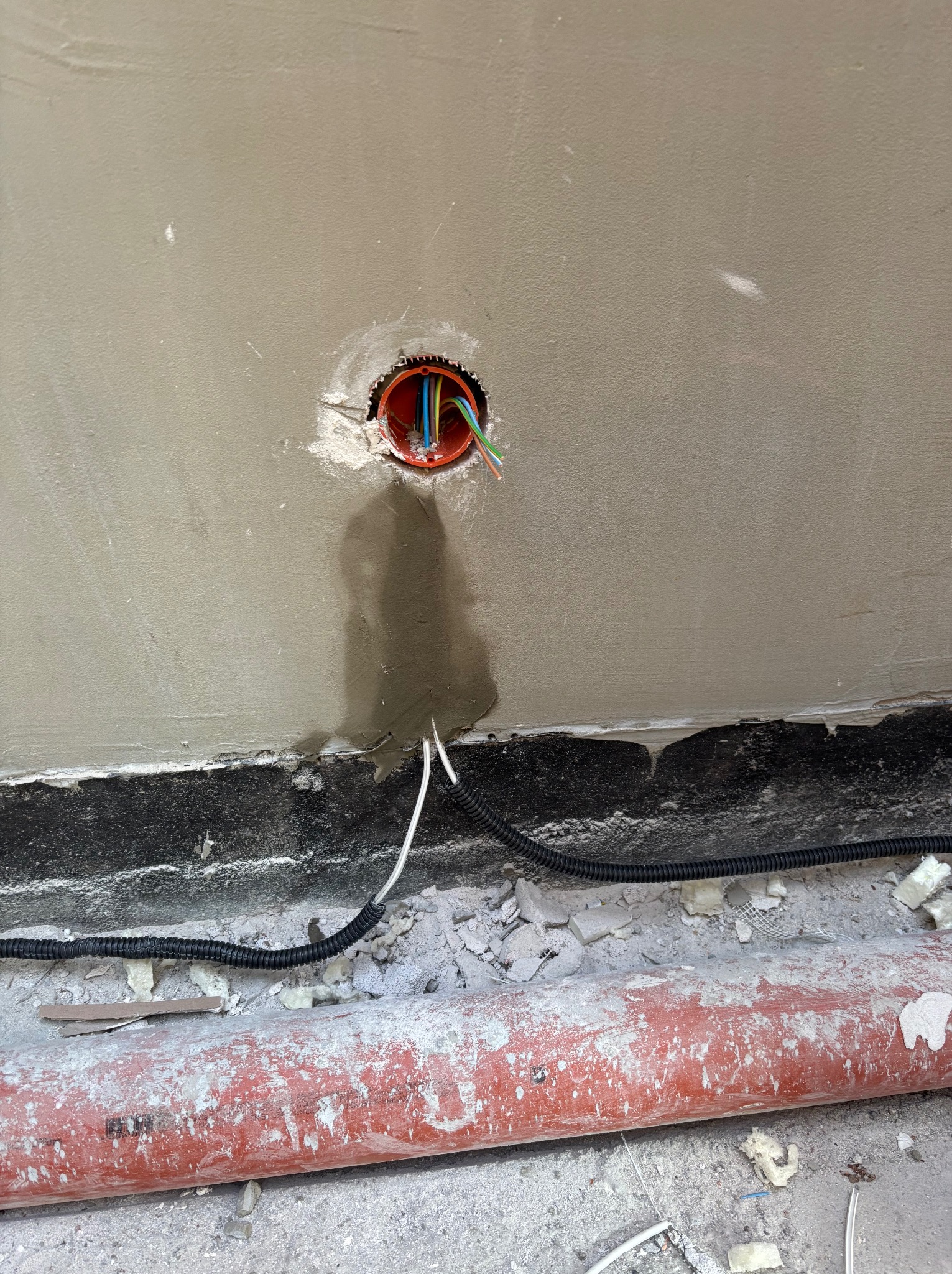 Pomarańczowa puszka instalacyjna z kolorowymi przewodami w szarej ścianie, widoczne kable i rura w podłodze. Etap prac instalacyjnych w budynku.