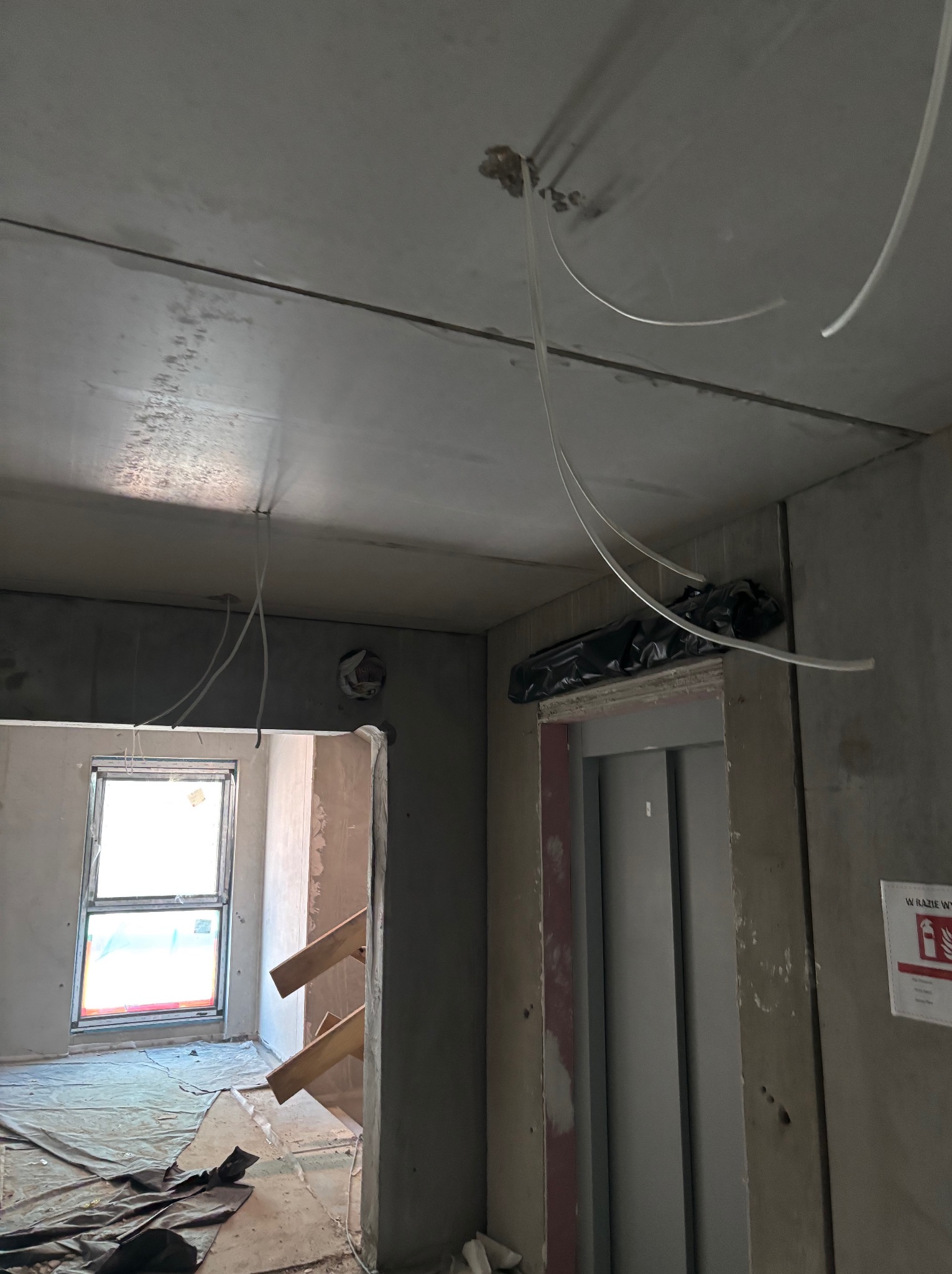 Surowy strop z wystającymi przewodami elektrycznymi w nowym budynku, widoczne okno i winda, stan deweloperski.