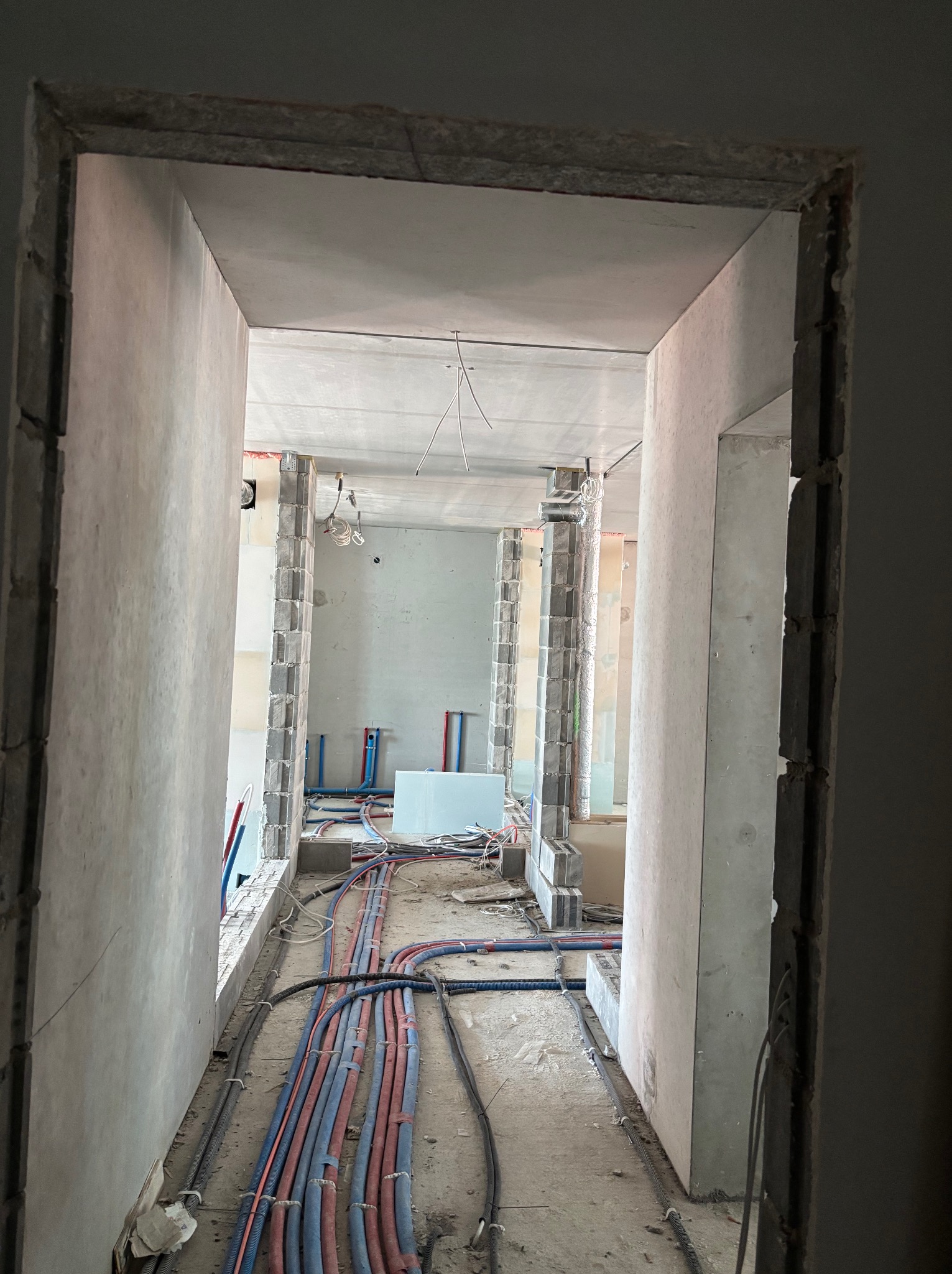 Wnętrze w trakcie remontu: widok przez surową futrynę na podłogę z licznymi przewodami elektrycznymi i rurami instalacyjnymi, ściany w stanie surowym, widoczne elementy instalacji.