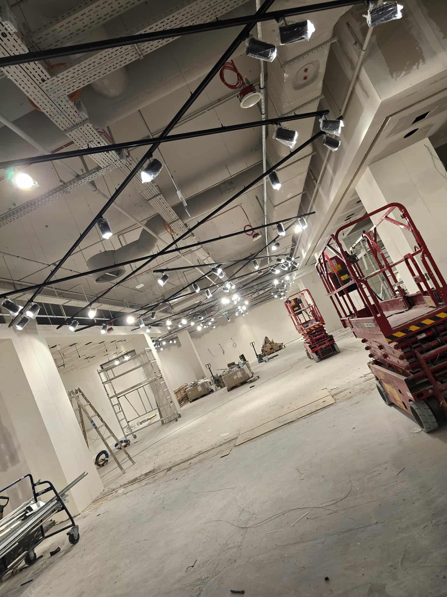 Montaż oświetlenia w nowym sklepie: sufit z instalacją, czarne szyny z reflektorami w folii, podnośniki koszowe, drabina. Wnętrze w trakcie wykończenia.