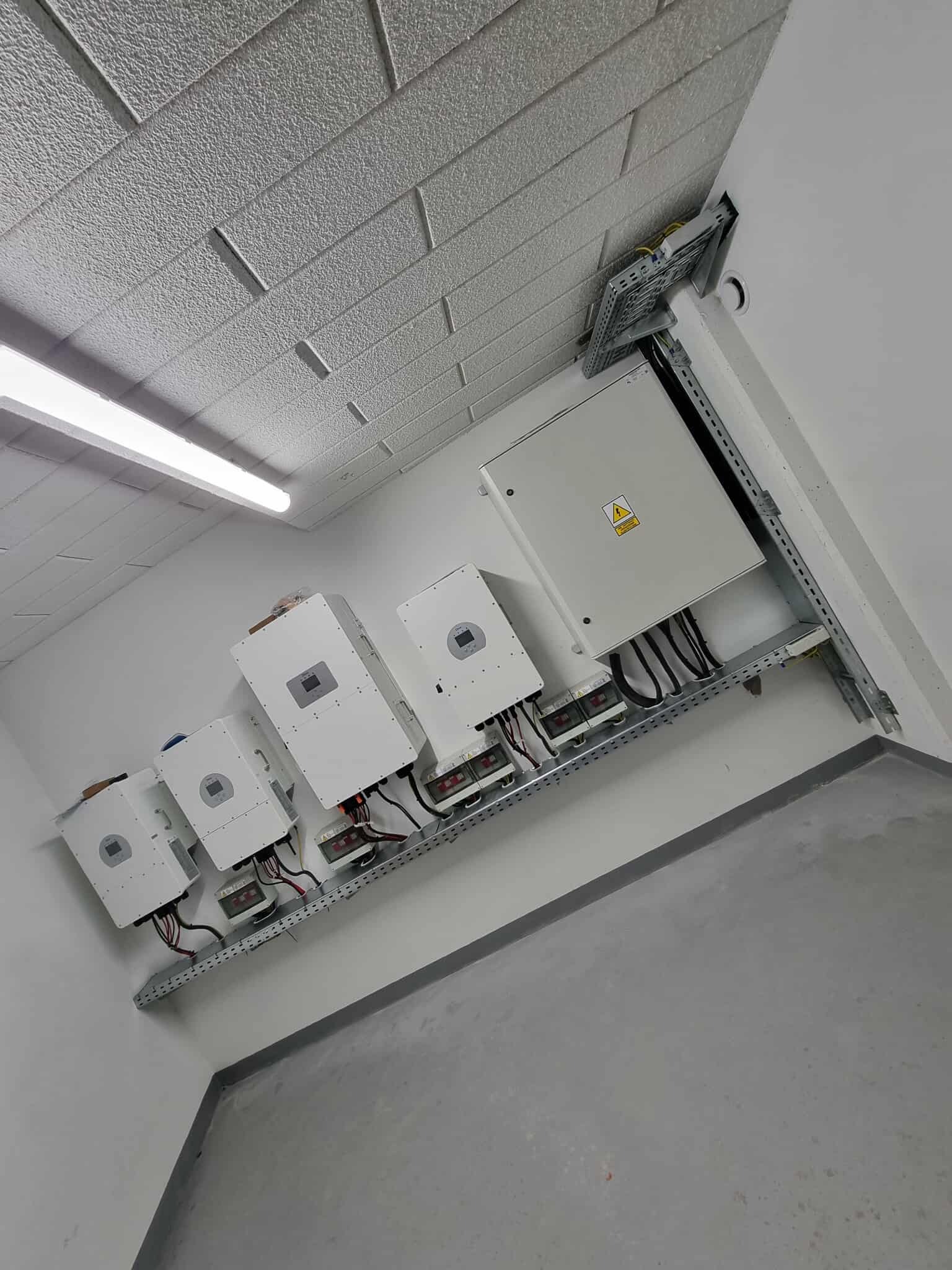 Rząd białych falowników solarnych zamontowanych na ścianie, z widocznymi kablami i skrzynką elektryczną z symbolem ostrzegawczym.