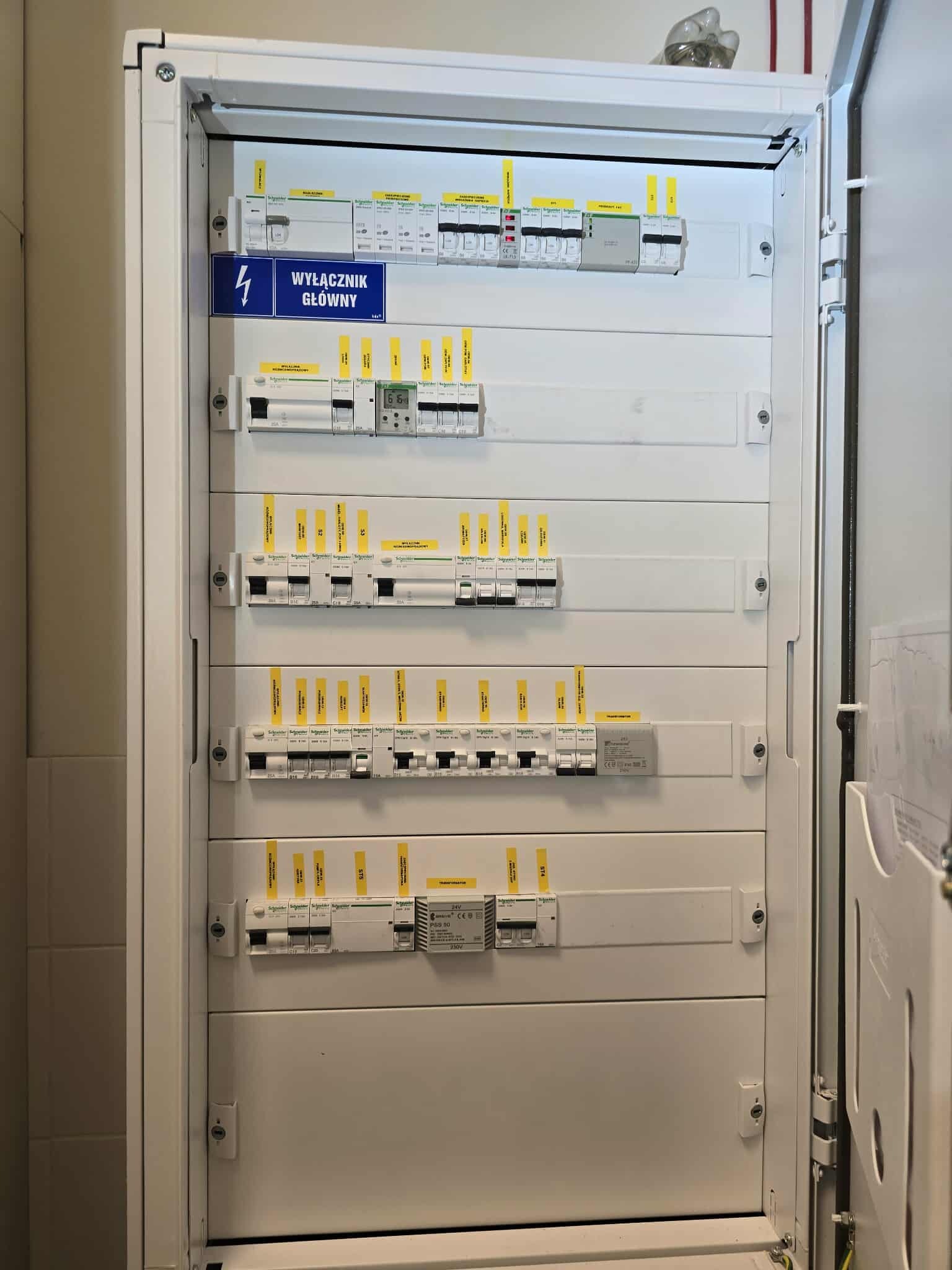 Rozdzielnica elektryczna w białej obudowie, z widocznymi bezpiecznikami i wyłącznikiem głównym oznaczonym niebieską etykietą. Widoczne liczne żółte oznaczenia na elementach.