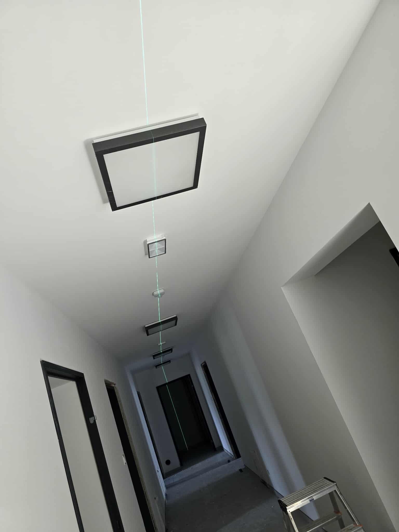 Montaż oświetlenia sufitowego w korytarzu z użyciem lasera, widoczne równo rozmieszczone lampy i drabina. Białe ściany i ciemne ramy drzwi.