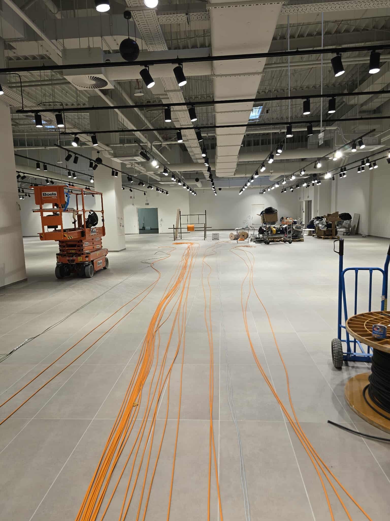 Rozciągnięte kable na podłodze w przestronnym pomieszczeniu z podnośnikiem i rozstawionym oświetleniem sufitowym. Etap instalacji elektrycznej w obiekcie komercyjnym.