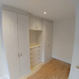 Jasna zabudowa szafy wnękowej z drewna z szufladami i szafkami wiszącymi, na podłodze widoczne ślady po remoncie.