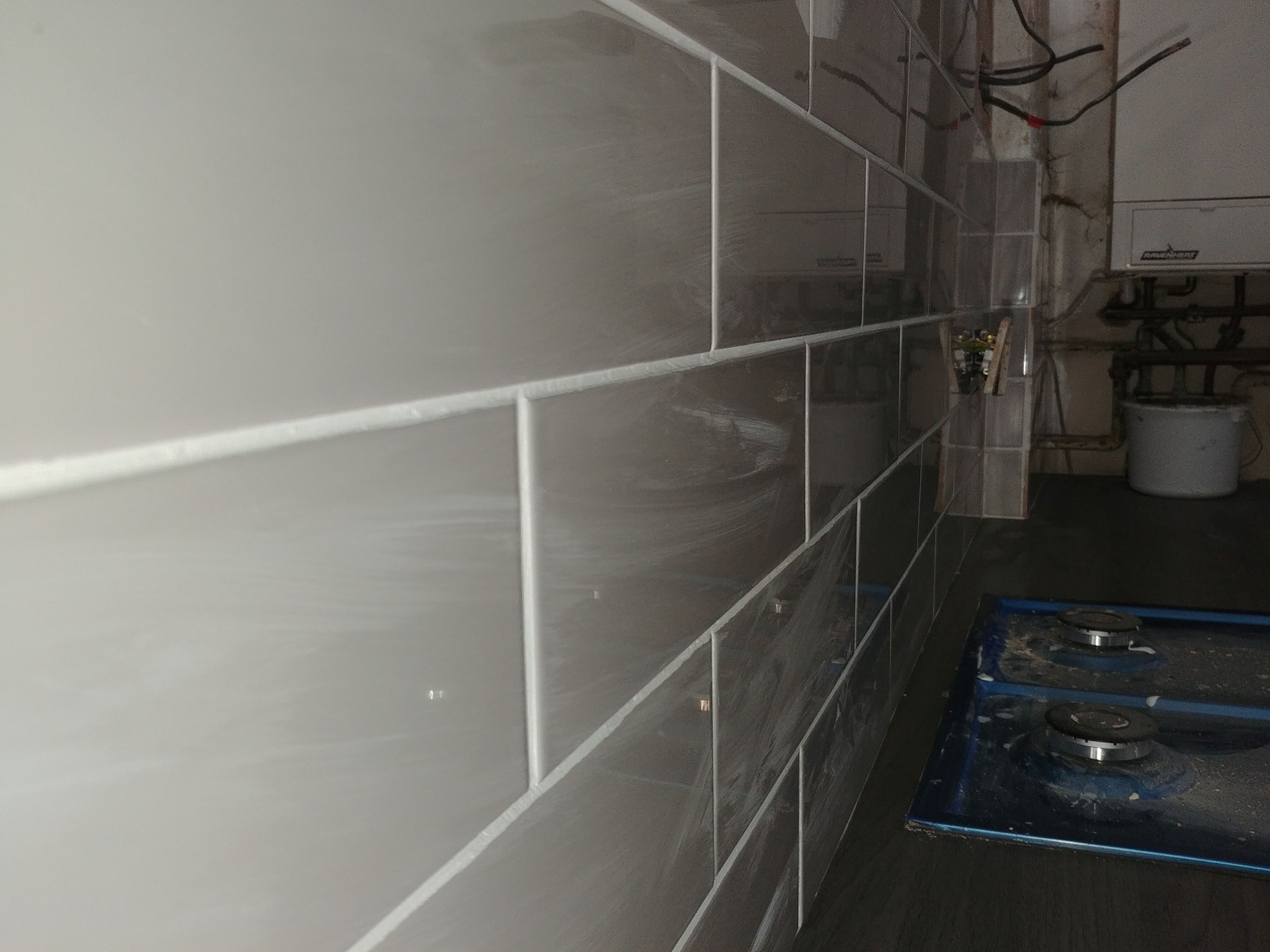 Świeżo ułożone, błyszczące, szare płytki na ścianie w kuchni, z widoczną kuchenką gazową i instalacją hydrauliczną w tle.