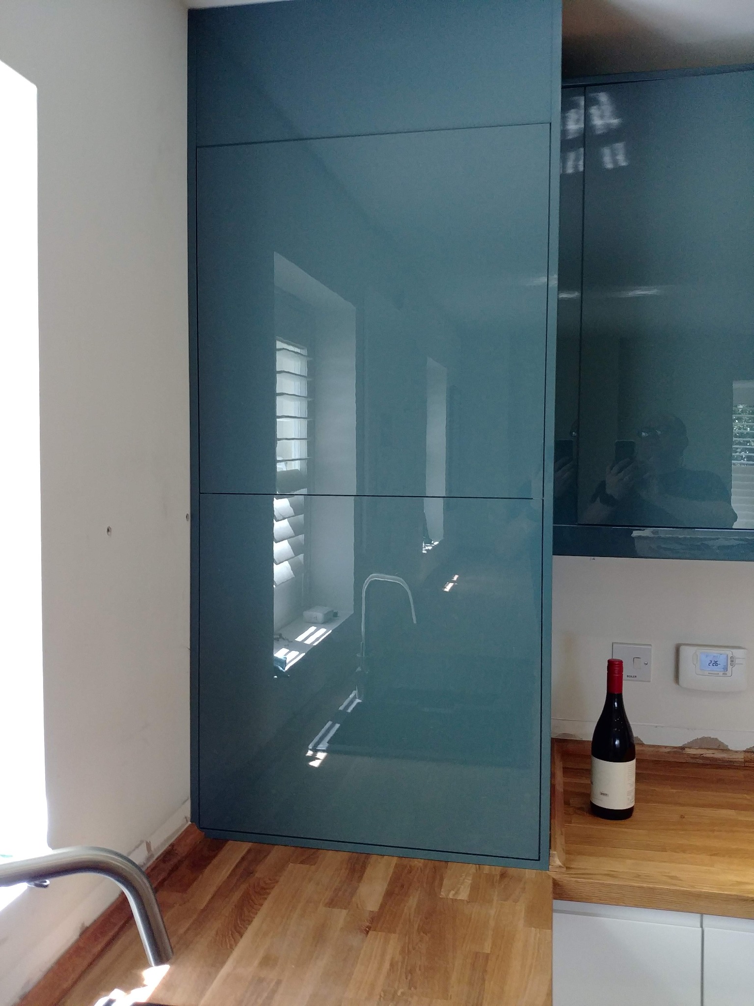 Błękitne, lakierowane fronty szafek kuchennych w Łodzi, z odbiciem okna i osoby robiącej zdjęcie, uzupełnione drewnianym blatem i butelką wina.