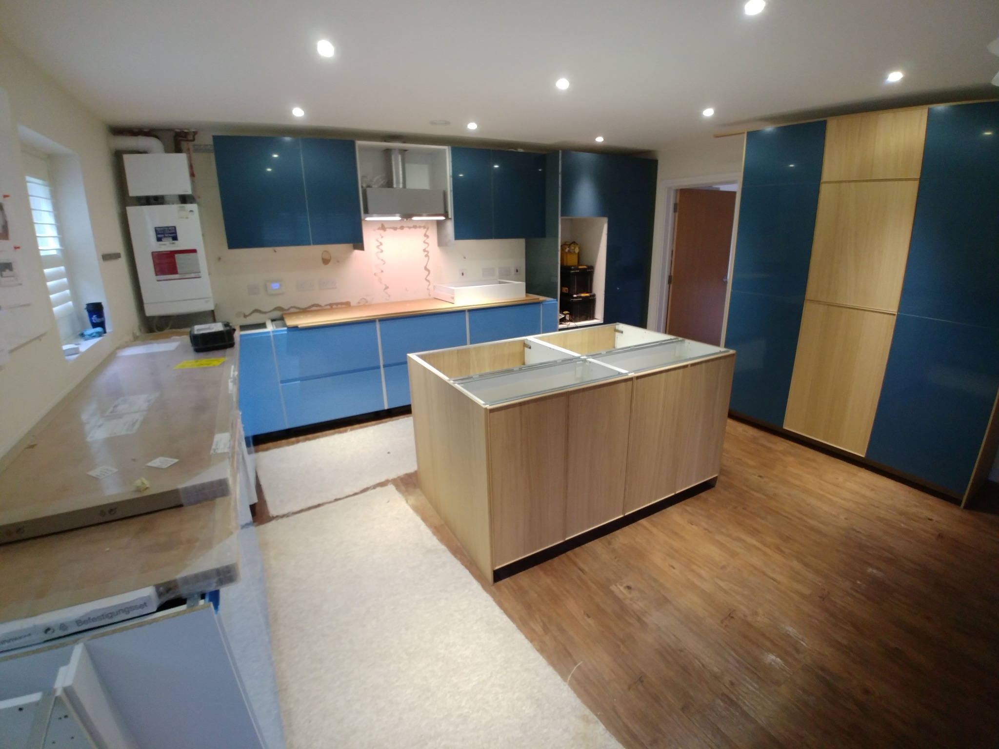 Wnętrze kuchni w trakcie montażu: niebieskie szafki, drewniany blat i wyspa kuchenna bez wykończenia, widoczny piec grzewczy na ścianie, podłoga częściowo zabezpieczona białym materiałem.
