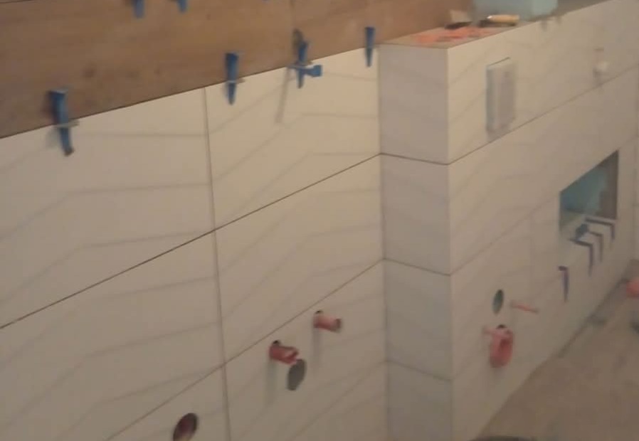 Ściana łazienki w trakcie układania białych płytek z delikatnym wzorem, użyte niebieskie klipsy poziomujące, widoczne otwory na instalacje wodne i elektryczne.