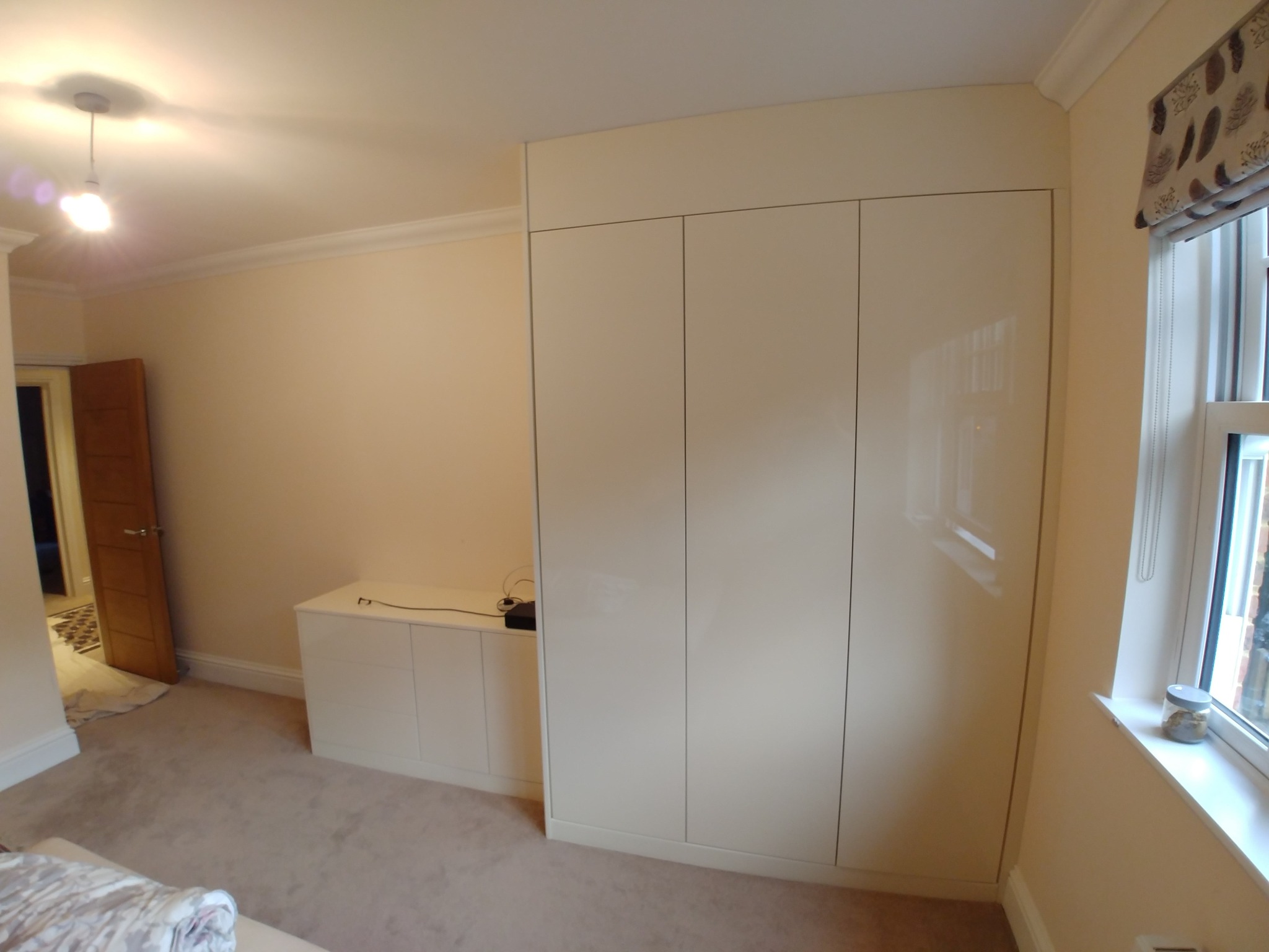 Wnętrze sypialni z jasną, zabudowaną szafą z czterema drzwiami na wysoki połysk, dopasowaną szafką RTV, oknem z roletą rzymską i dywanem w odcieniach beżu.