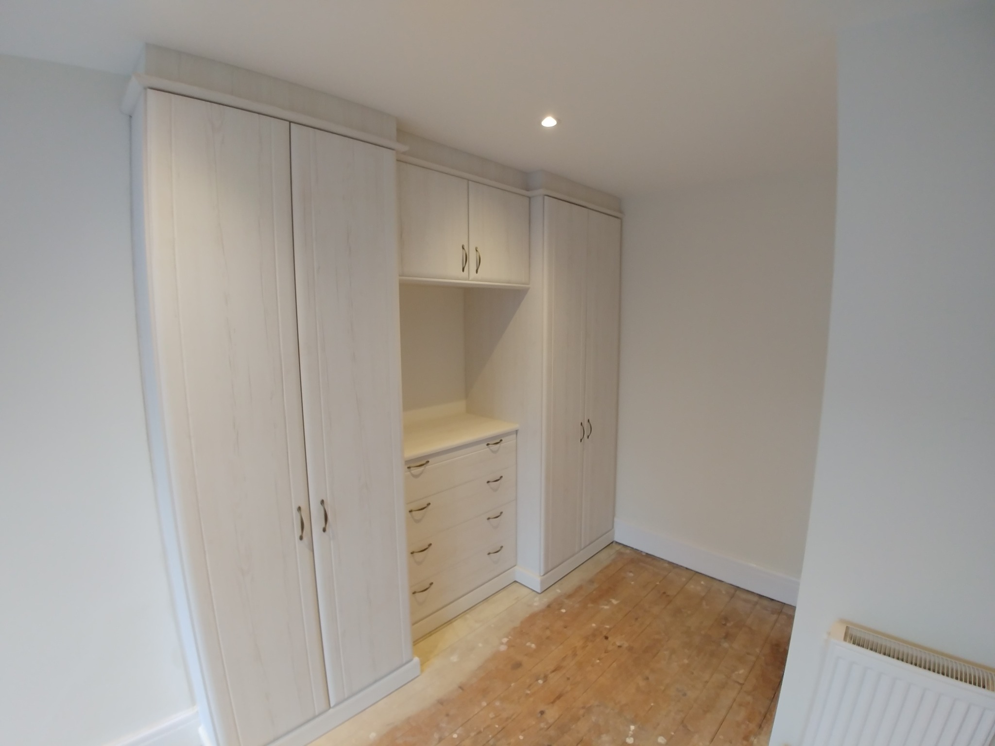 Jasna zabudowa szafy wnękowej z drewna z szufladami i szafkami wiszącymi, na podłodze widoczne ślady po remoncie.