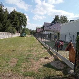 Dębowski Paweł Wyrób i Sprzedaż Siatki Ogrodzeniowej - Nowoczesne ogrodzenie panelowe z podmurówką betonową wzdłuż posesji, z zaparkowanym skuterem i budą dla psa w pobliżu, widok na domy i zieleń w tle.