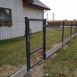 Dębowski Paweł Wyrób i Sprzedaż Siatki Ogrodzeniowej - Nowoczesne ogrodzenie z paneli w kolorze antracytowym z otwartą furtką, zamontowane na betonowej podmurówce imitującej cegłę, widok na dom z oknem i kwiatami w doniczce.