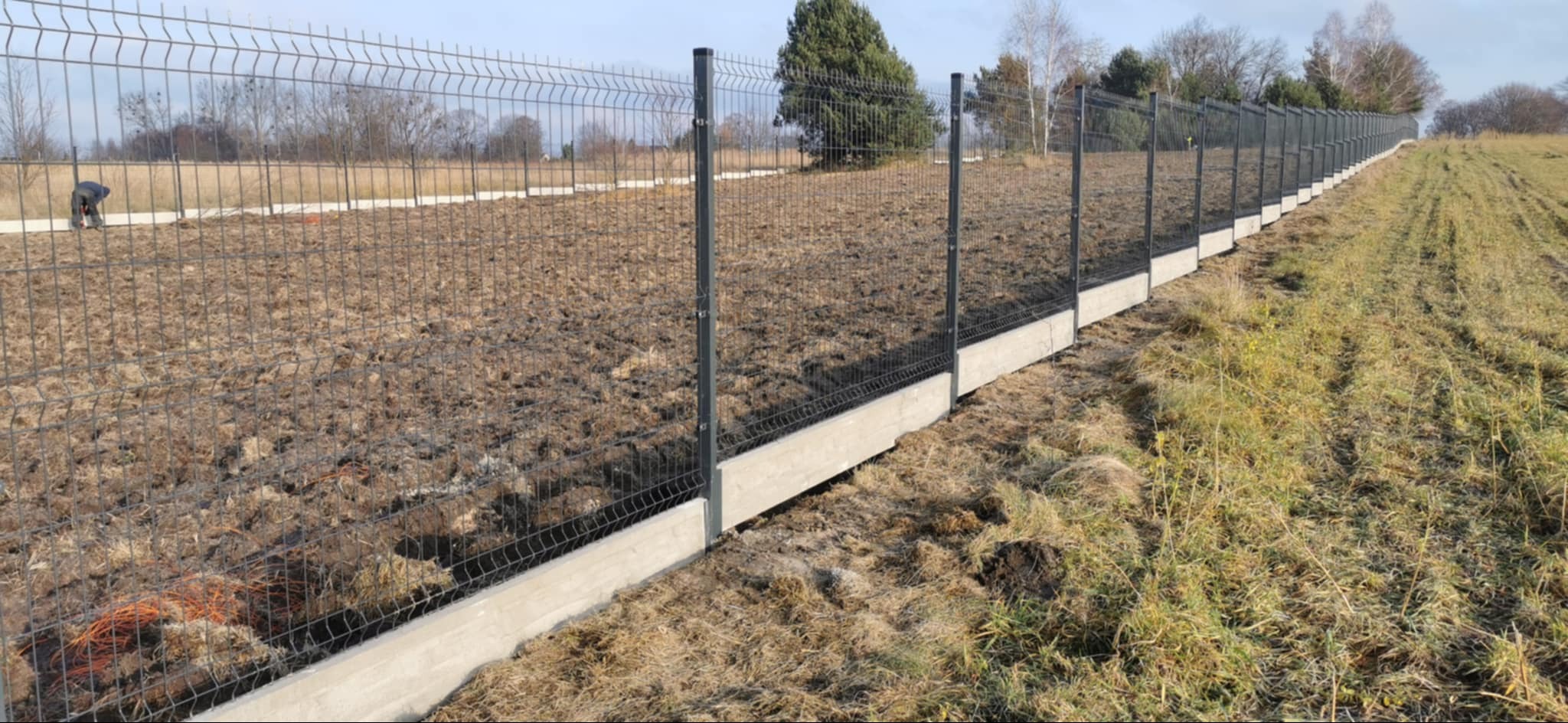 Długi, szary system ogrodzeniowy z paneli na betonowej podmurówce oddzielający pole uprawne od łąki z widocznym pracownikiem w oddali.