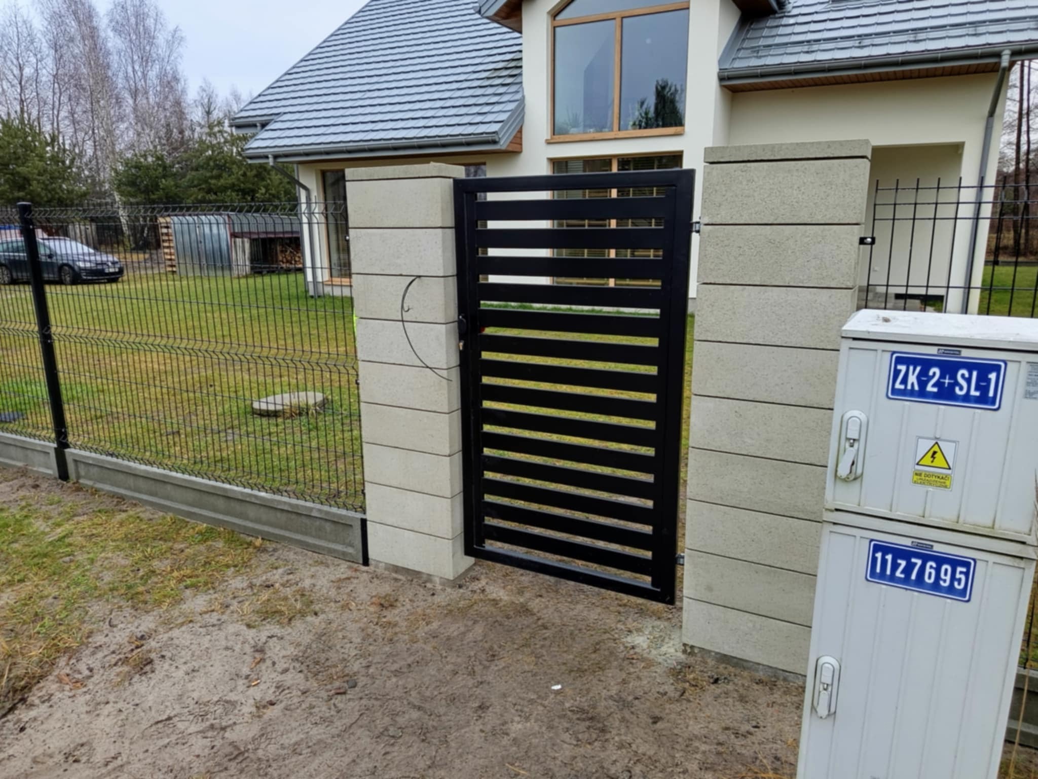 Nowoczesna, pozioma brama metalowa w kolorze czarnym, zamontowana między dwoma betonowymi słupkami, z przyległą siatką ogrodzeniową na betonowej podmurówce, na tle domu z szarym dachem i trawnika.