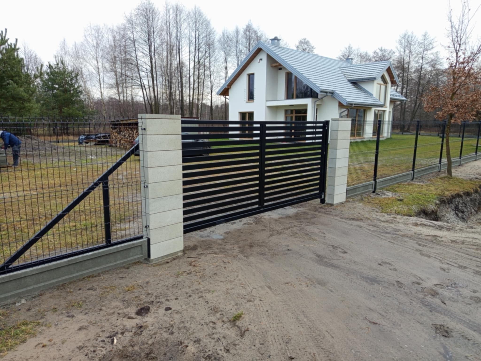 Nowoczesna posesja z czarną, poziomą bramą przesuwną i ogrodzeniem z siatki, widoczne jasne słupki z imitacją kamienia i dom w tle.