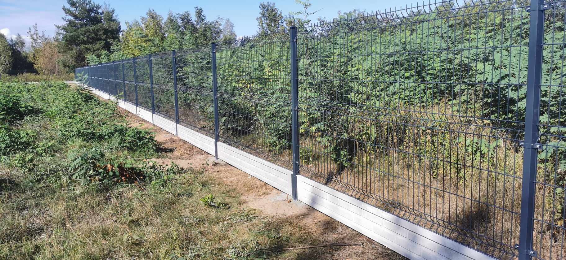 Długi, szary panelowy płot z betonową podmurówką, otoczony zielenią drzew i trawy.