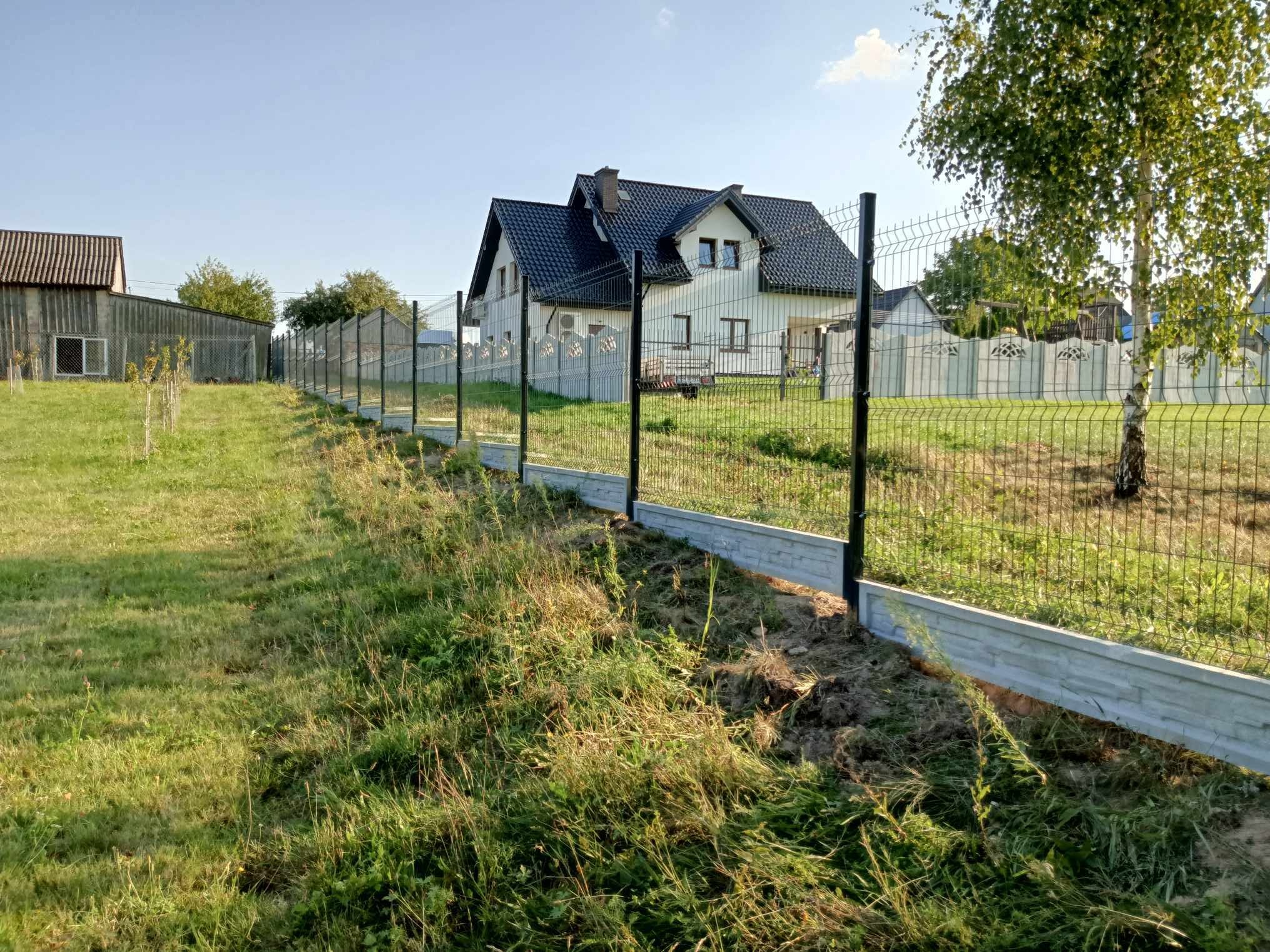 Nowo postawione ogrodzenie z siatki z podmurówką betonową, widok wzdłuż linii ogrodzenia w kierunku domu z czarnym dachem i białymi ścianami, trawa częściowo wykoszona.