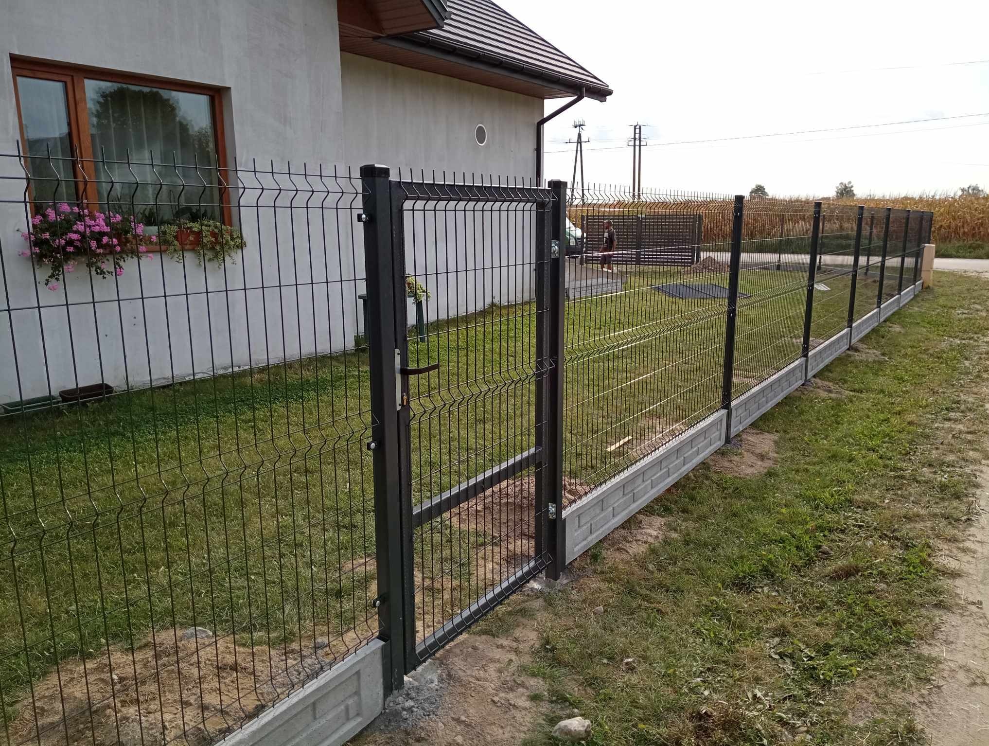 Nowoczesne ogrodzenie z paneli w kolorze antracytowym z otwartą furtką, zamontowane na betonowej podmurówce imitującej cegłę, widok na dom z oknem i kwiatami w doniczce.