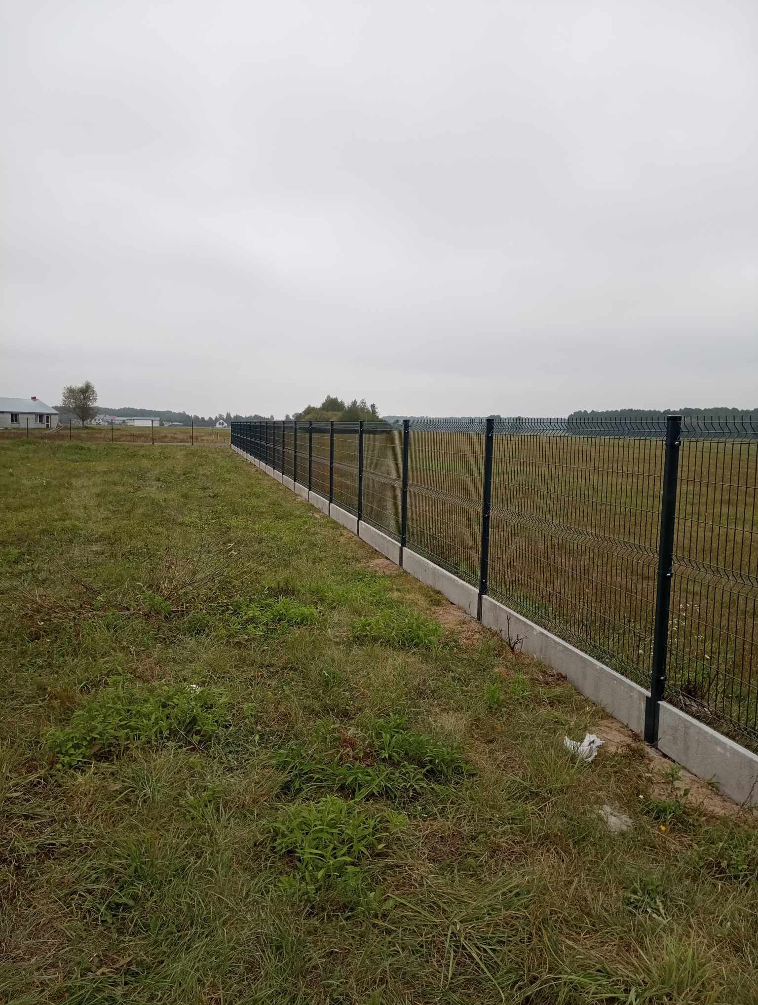 Nowoczesne ogrodzenie z paneli z podmurówką betonową, widok na rozległe pole i zabudowania w tle, pochmurny dzień.