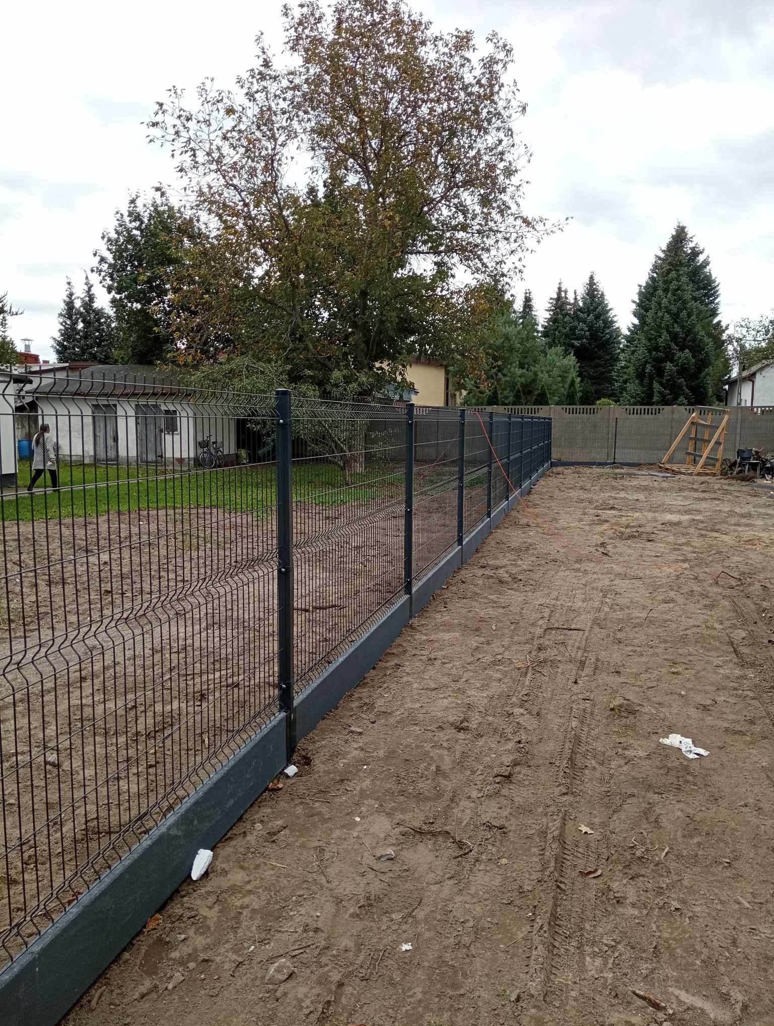 Nowo postawione ogrodzenie z siatki z podmurówką betonową, widoczne ślady opon na ziemi, w tle budynek gospodarczy i drzewa.