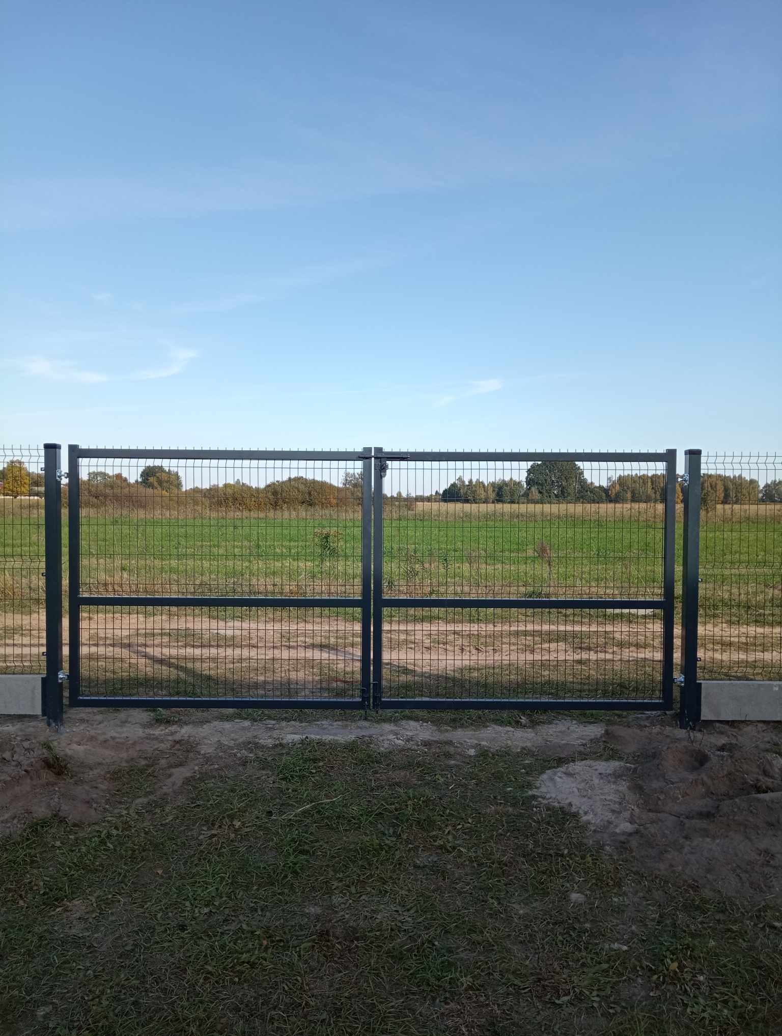 Zamknięta, dwuskrzydłowa brama z siatki panelowej, osadzona na betonowych słupkach, na tle łąki i błękitnego nieba.