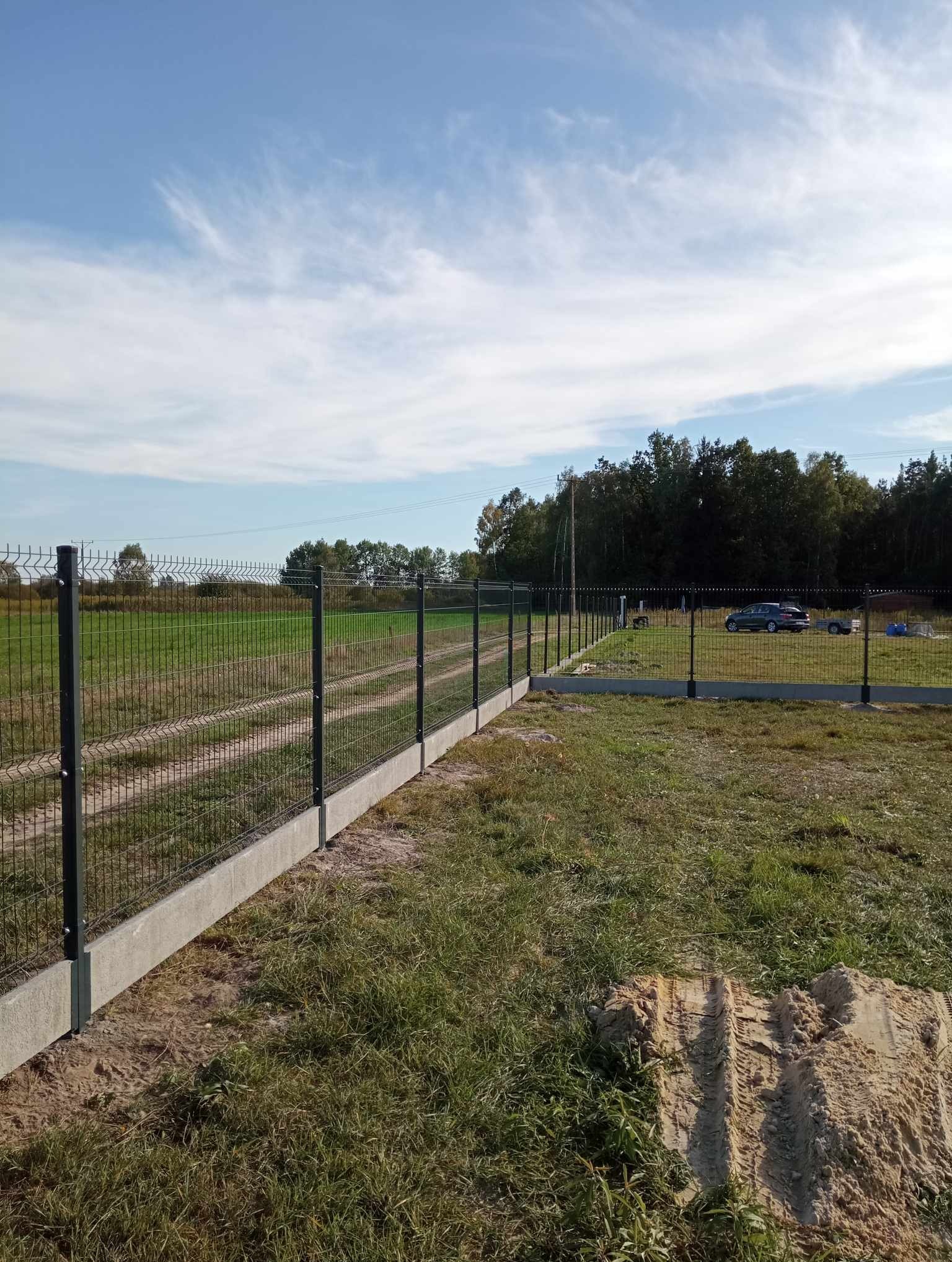 Zmontowane ogrodzenie panelowe na betonowej podmurówce, widok na teren z samochodem i przyczepą, błękitne niebo z chmurami w tle.
