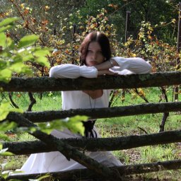 Olaf Chalecki - Młoda kobieta w białej sukni, opierająca się o stary, omszały drewniany płot na tle winnicy.