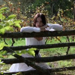 Młoda kobieta w białej sukni, opierająca się o stary, omszały drewniany płot na tle winnicy.